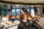 Ein Mikrofon, im Hintergrund sind sitzende Personen zu sehen.