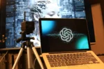 Ein Macbook, auf dem das OpenAI-Logo zu sehen ist. Im Hintergrund eine Grafik eines Roboters.